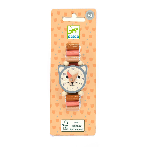 A DJECO wooden cat-shaped watch on a peach-colored backing card. The watch features a gray cat face with a white clock face, pink hands, and a brown and pink wooden link strap with white wooden beads.