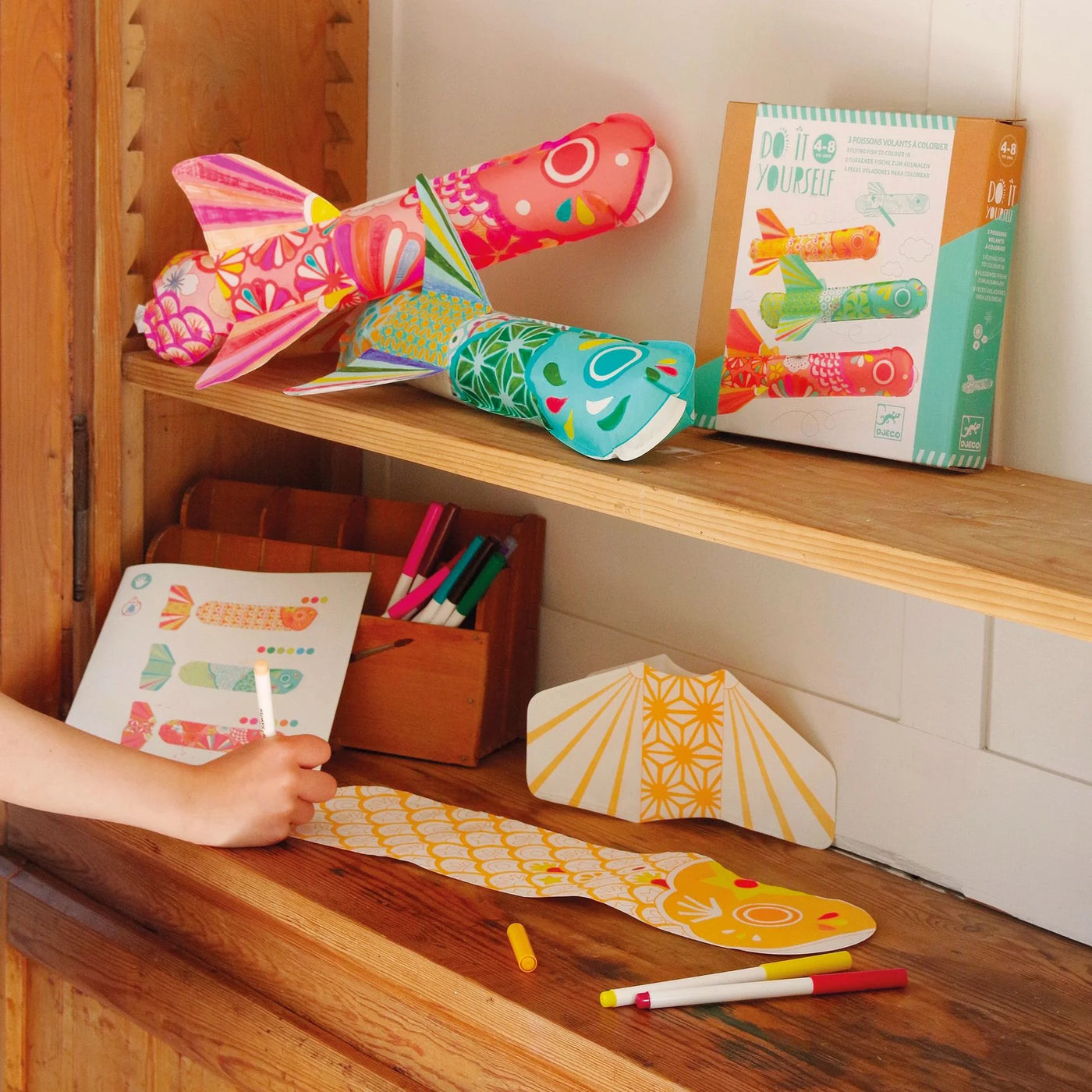 Colourful fish-shaped toys on a wooden shelf with a child's hand interacting with a paper craft.