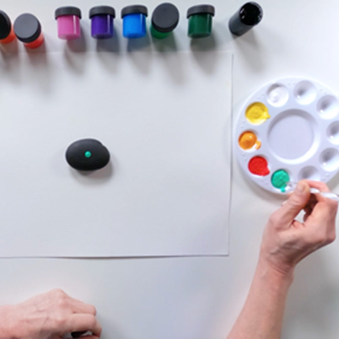 Person painting a rock with a paintbrush next to a palette of colors on a white surface.
